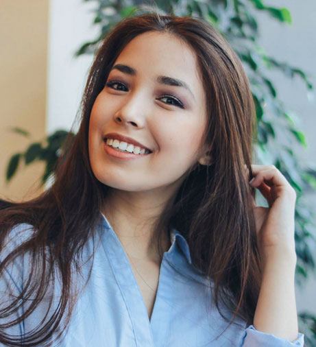 Young woman smiling at camera in light blue shirt, indoor setting with plants.