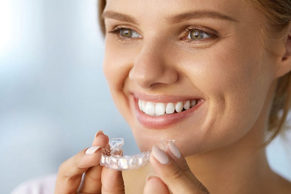 Woman smiling while holding clear dental aligner tray against blurred background.