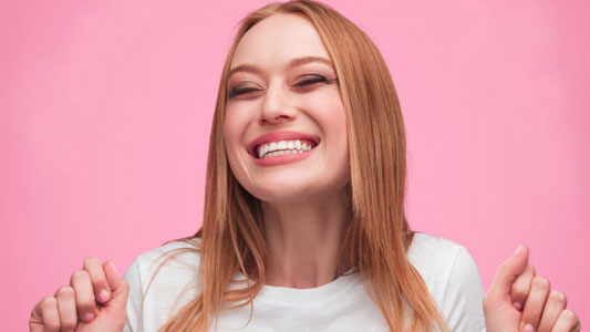 Smiling woman with reddish-blonde hair against pink background, giving thumbs up gesture.