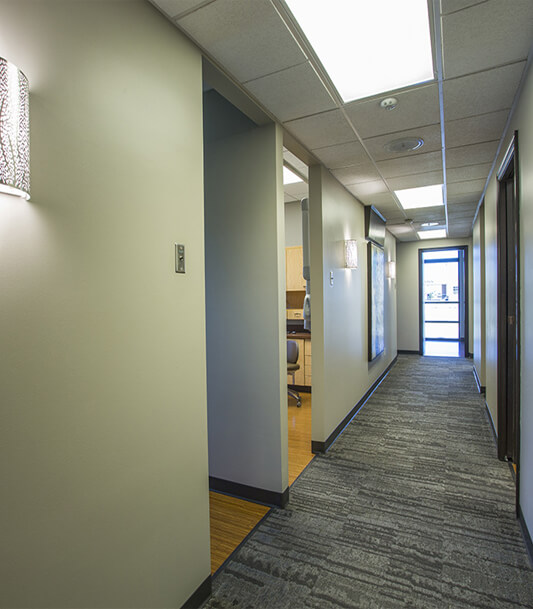 Modern dental office hallway with light green walls, overhead lighting, and wooden door frames.