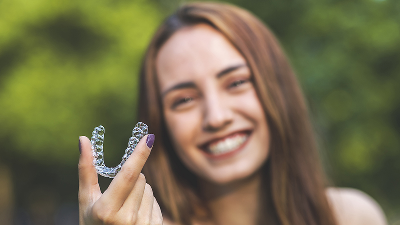 Young woman smiling outdoors making peace sign with fingers, green blurred background.
