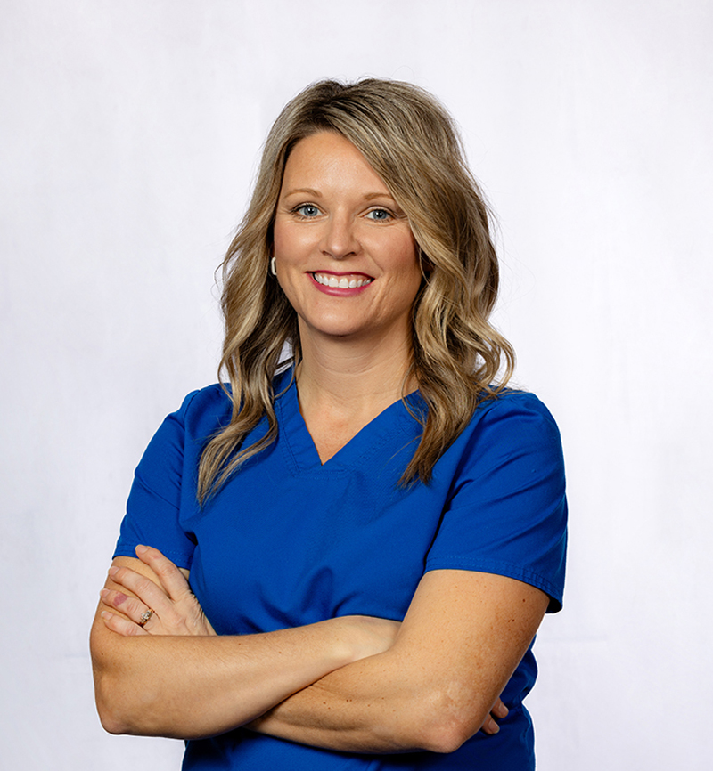 Female dental professional in blue scrubs with arms crossed, smiling at camera against white background.