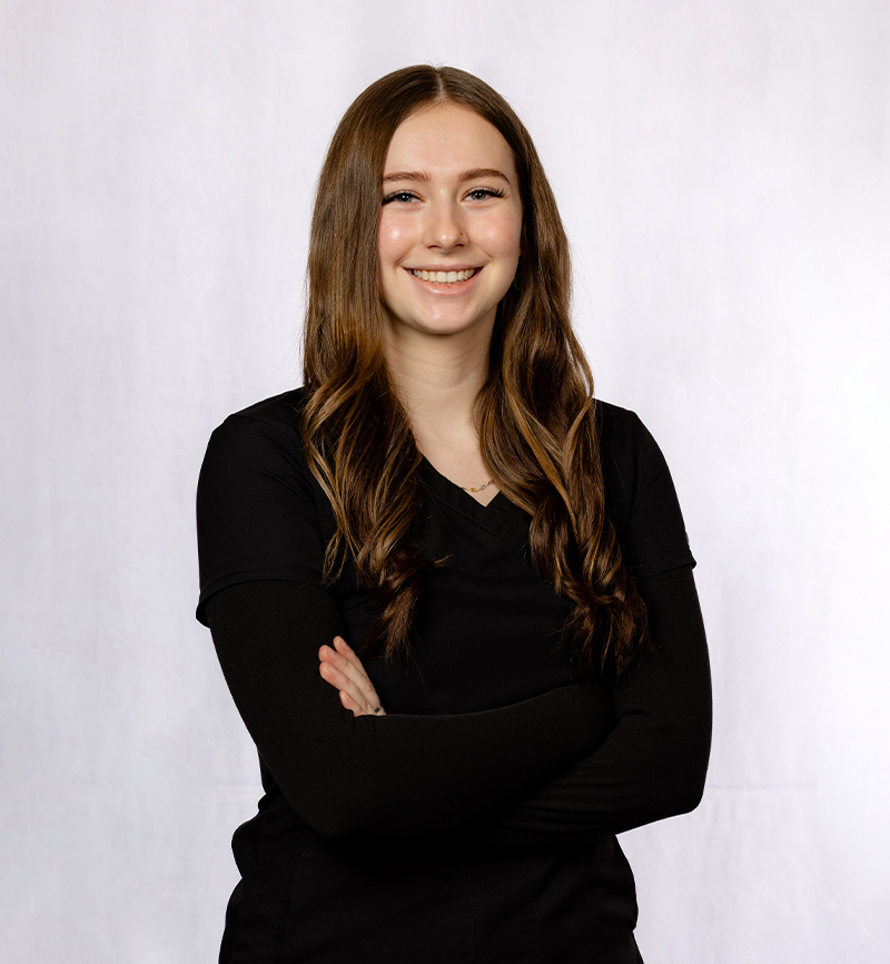 Professional headshot of smiling woman with long brown hair, arms crossed, wearing black blazer against white background.