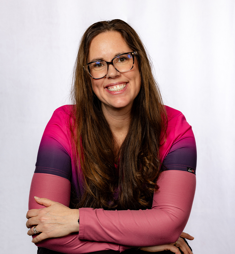 Woman wearing glasses and pink striped shirt smiles at camera in professional portrait setting.