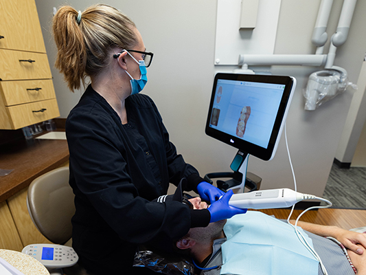 Dental hygienist wearing blue gloves and mask examines patient using intraoral camera in clinical setting.