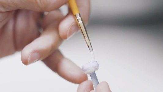 Hand holding paintbrush applying white dental material to tooth model in clinical setting.