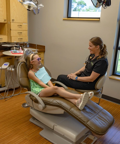 Pediatric dental patient reclines in treatment chair while dentist examines teeth in modern dental office.