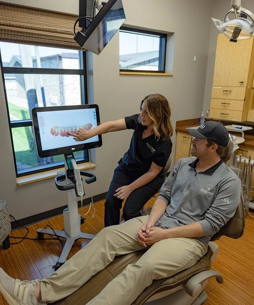 Dental professional pointing at monitor while patient sits in treatment room during consultation.