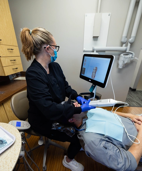 Dental professional wearing blue gloves and glasses working at computer in clinical office setting.